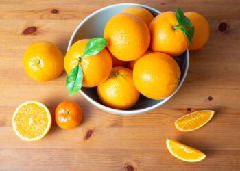 Top view of big bowl with bright oranges standing on wooden desk. Mandarin and cut tropical fruit laying on table. Studio shot. Nutrition and vegetarian concept